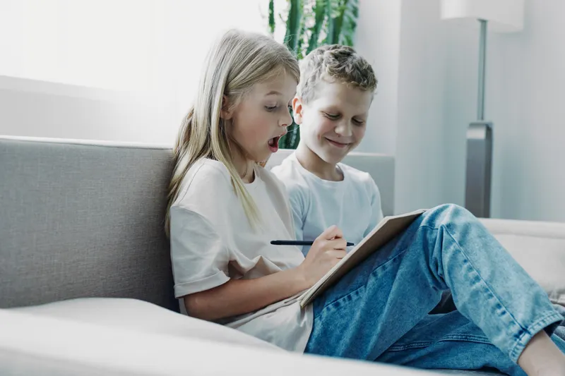 Two children sitting on a couch, girl surprised while writing on a notebook, boy smiling nearby. Indoor scene with natural light and casual clothing, concept of learning and creativity.