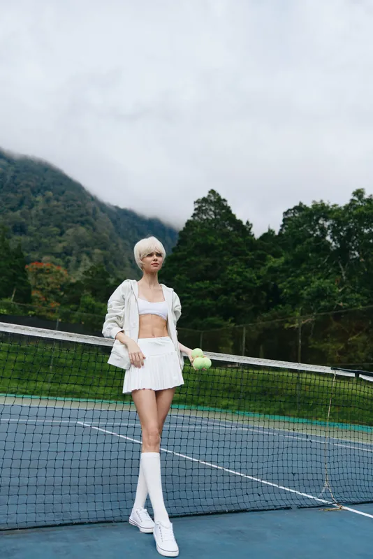 Confident female tennis player wearing a stylish white sports outfit, holding a tennis ball, standing on a court surrounded by nature, perfect for sports and fitness themes