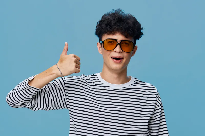 Young man wearing striped shirt and orange sunglasses shows thumbs up sign with happy expression, isolated on blue background in casual style and cheerful mood.
