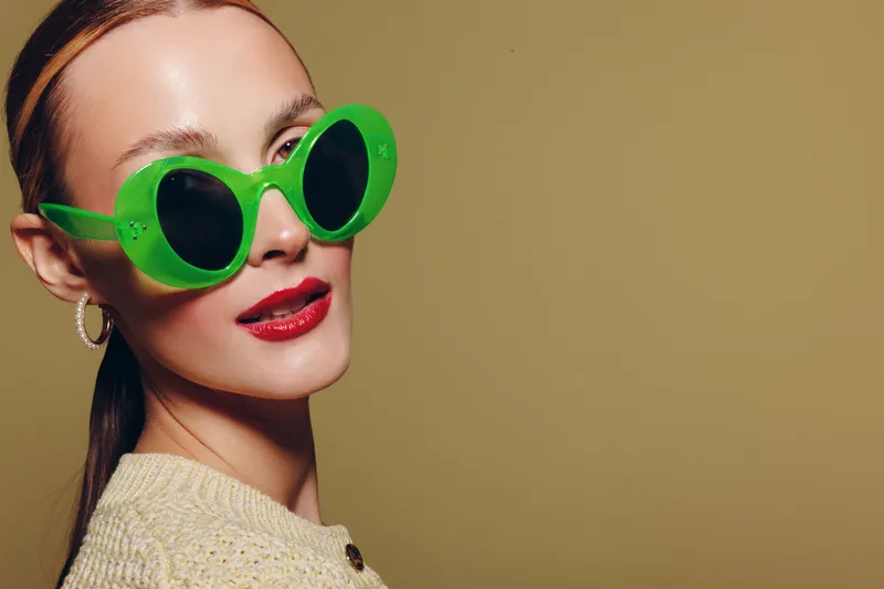 Stylish woman with green sunglasses and red lipstick poses against beige background. Fashion portrait with trendy accessories and confident expression. Concept of modern style and beauty.