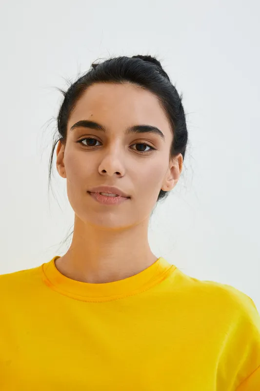 young woman portrait with dark hair and serene expression wearing bright yellow shirt against plain white background, natural light emphasizes calm and simplicity