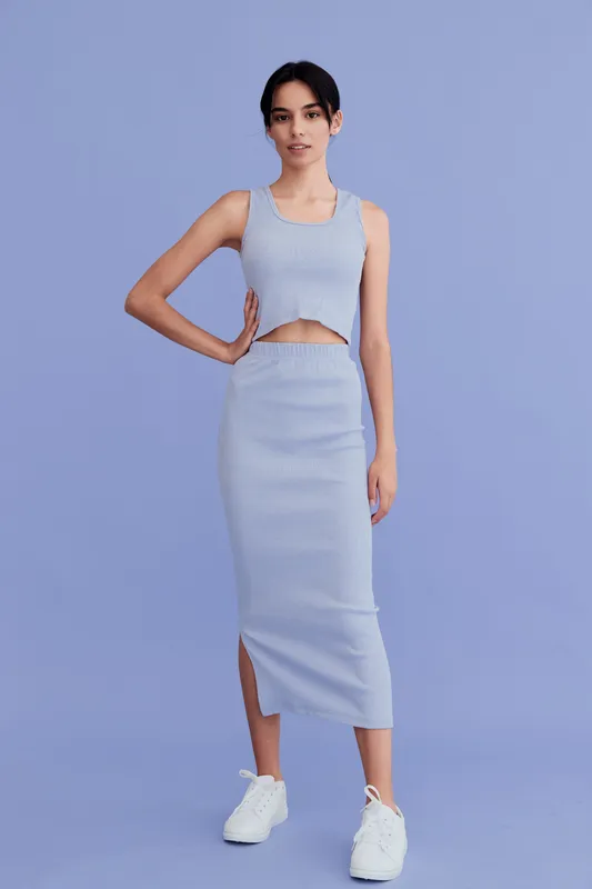 young woman in blue matching crop top and skirt, posing with hand on hip, wearing white sneakers, standing on solid periwinkle background, casual summer outfit