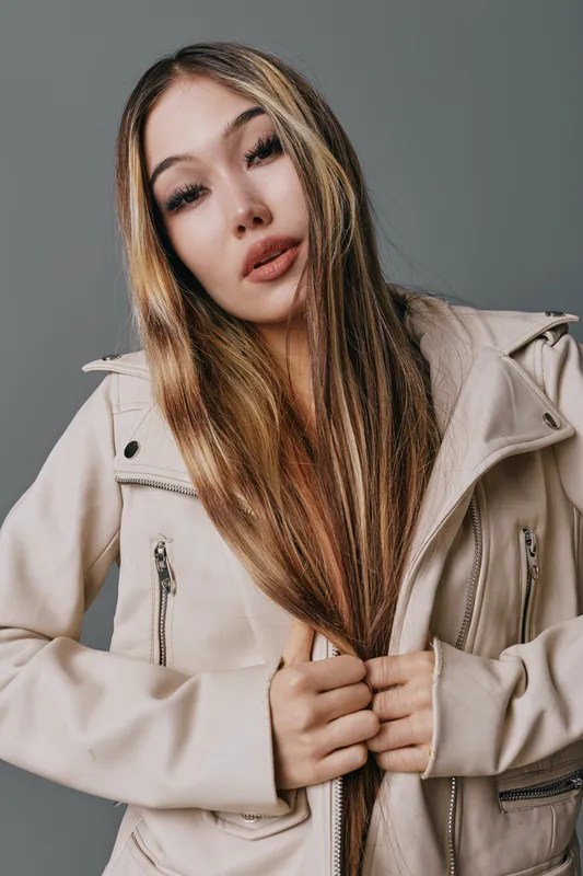 Young woman with long straight hair wearing light beige jacket standing against plain gray wall with serious expression holding jacket zipper with both hands.