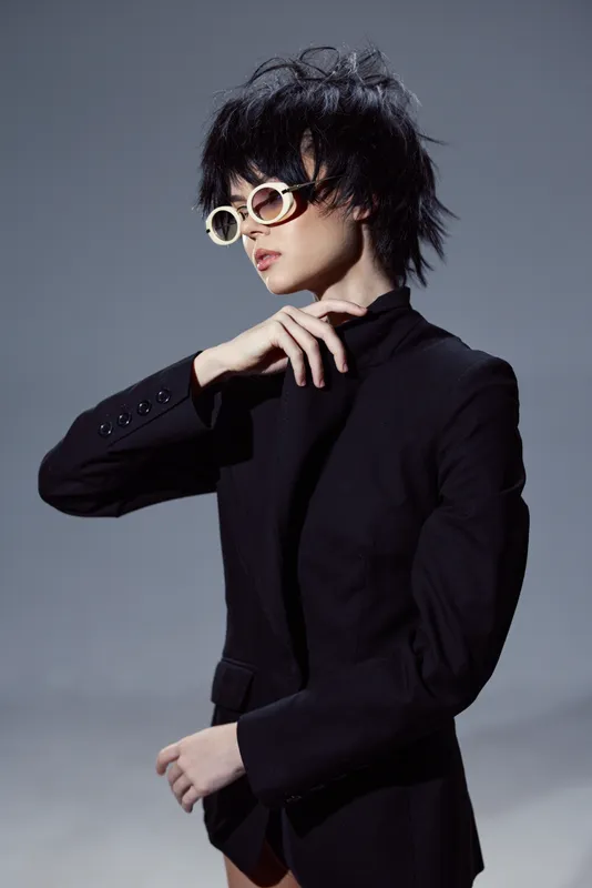 Stylish woman in black blazer wearing round glasses against gray background with modern hairstyle and thoughtful expression