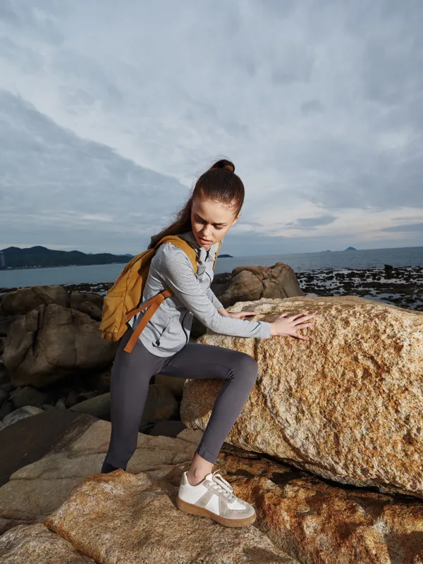 Young female hiker climbing on rocky terrain by the sea, wearing activewear and a backpack, showcasing determination and adventure spirit against a cloudy sky