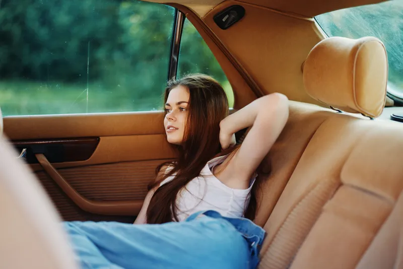 Young woman relaxing in a vintage car seat, wearing a casual outfit, expressing a thoughtful emotion and surrounded by a lush green background.