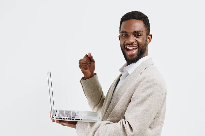 Smiling man with dark skin holding a silver laptop in hand, wearing a beige knitted cardigan and white shirt, looking at camera, happy expression, isolated on plain white background, studio portrait, 