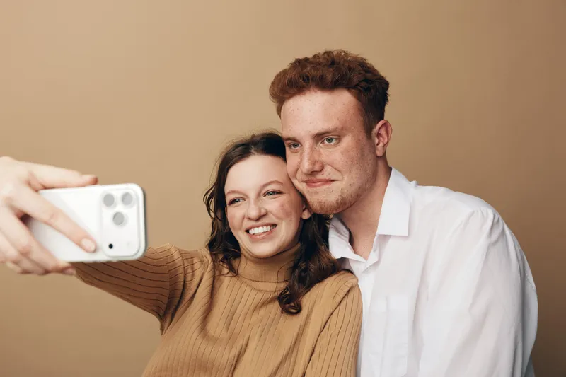 Smiling young couple takes a selfie together on a smartphone. Woman in brown sweater and man in white shirt pose closely on beige background. Concept of friendship and casual lifestyle.