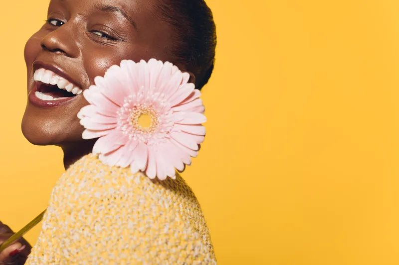 Happy smiling African woman with dark skin holds pink flower near face against yellow background. Concept for beauty, skincare, natural glow, and positive emotions in studio lighting.