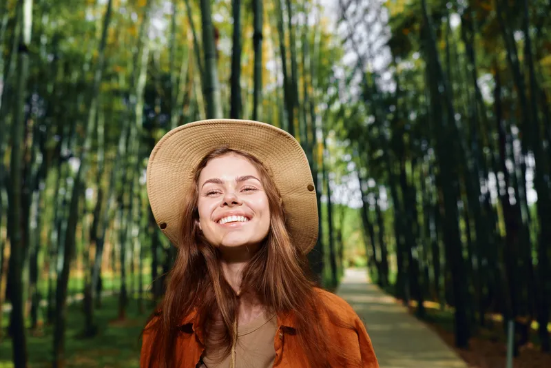 Woman with smile in straw hat on woodland path surrounded by tall trees, nature background and sunlight. Portrait of happy traveler in autumn jacket enjoying outdoor forest walk.