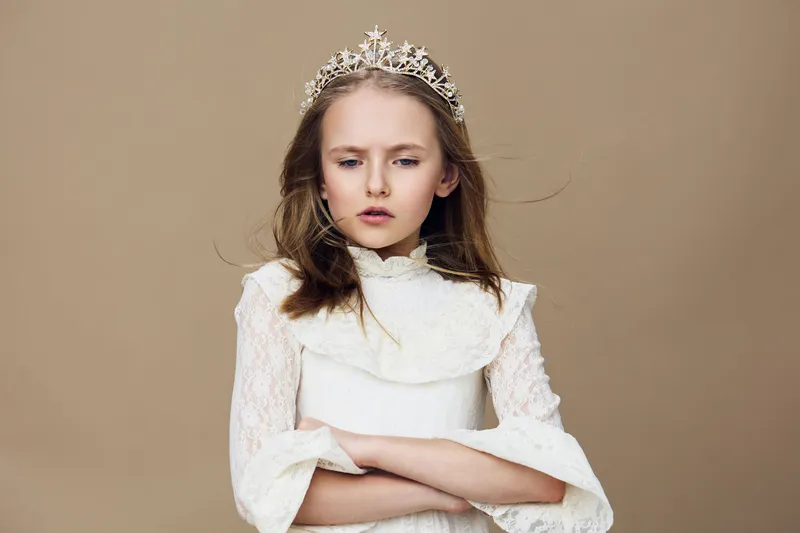 A young girl in a white dress with a crown, resembling royalty, expressing a serious emotion against a soft beige background for portrait photography