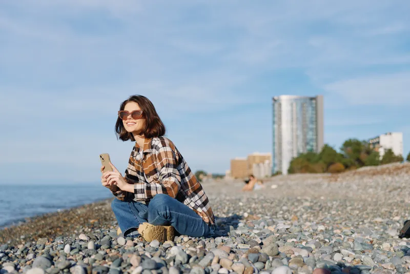Woman on a pebble beach wearing a plaid shirt and sunglasses, sitting by the sea while using a phone, with distant buildings and blue sky in the background.