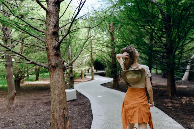 Woman walking on a winding pathway through park trees, back view of a fashionable outfit with orange skirt and heart top, casual summer stroll in green urban forest setting.
