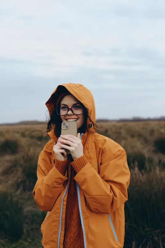 Woman lifestyle outdoors, smiling in orange hood coat, holding coffee mug, glasses visible, standing in brown field, cloudy sky background, warm comfort in cold weather