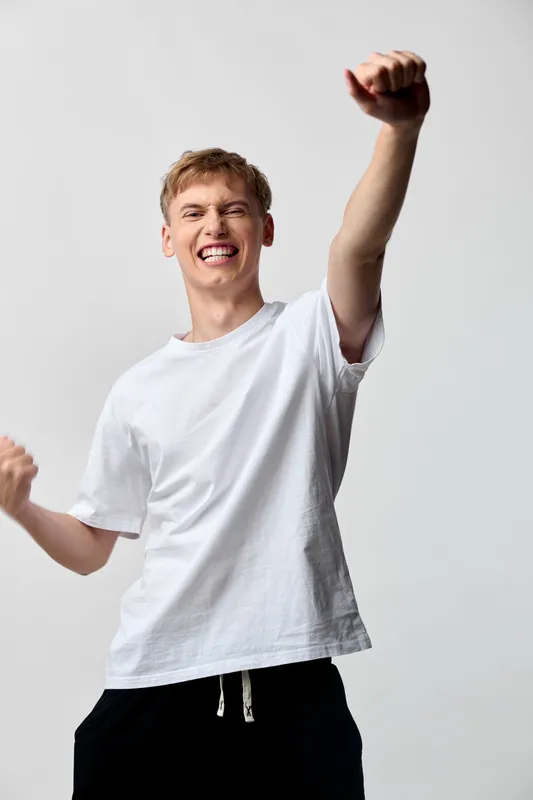 Celebration of achievement with a youthful man in a white tee raising his fist, expressing joy and energy, a moment of victory and happiness in a bright studio setting