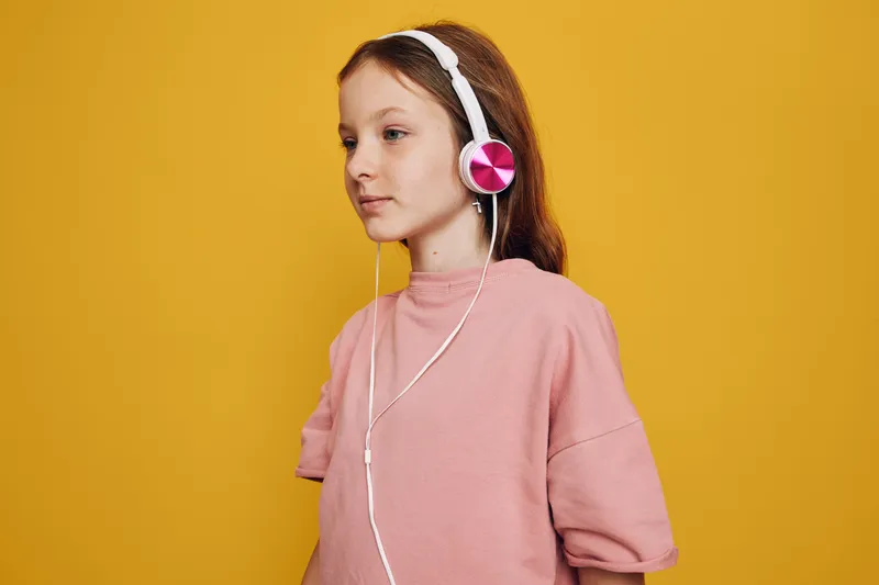 Young girl with long red hair listens to music using white headphones with pink accents. She wears a loose pink sweatshirt and stands against a solid yellow background. Concept for youth, technology,