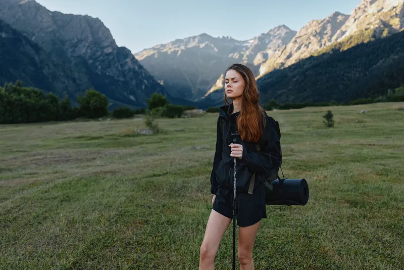 An attentive female photographer stands in a wide meadow beneath towering mountain peaks, holding a camera with a lens, ready to capture nature scenes with calm focus.