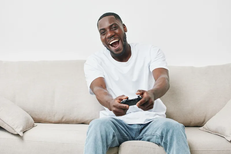Joyful young Black man enjoying video games in a comfortable living room, wearing a white t shirt and jeans, showcasing excitement and relaxation