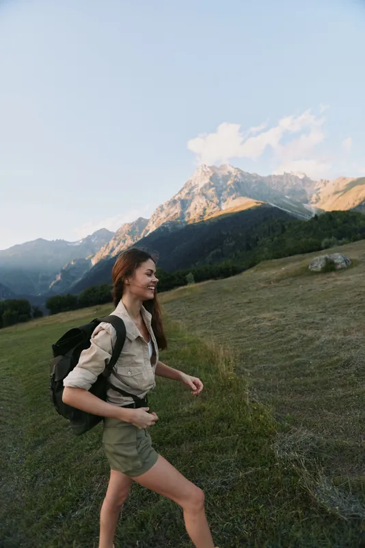 Woman hiker with backpack walks along a hillside trail in a mountain landscape, enjoying nature, fresh air, sunlight on peaks, casual outdoor attire, peaceful moments during an active lifestyle.