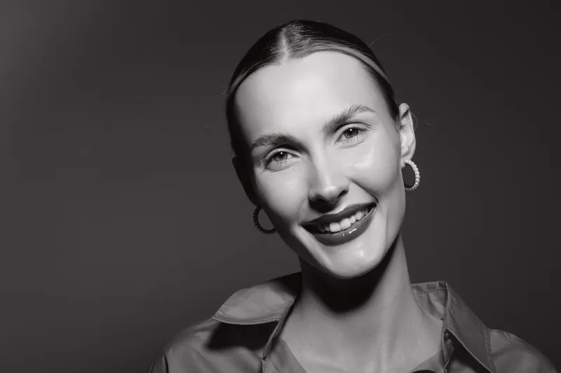 Close-up portrait of smiling Caucasian woman with fair skin and slicked-back hair wearing hoop earrings in black and white studio lighting.