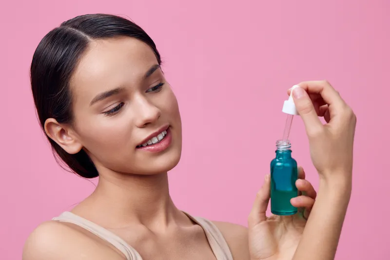 Young woman applying facial serum for skincare on pink background, expressing happiness and confidence in self care routine