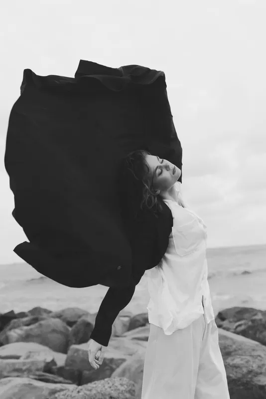 Wind catches a flowing scarf around a woman on a rocky beach with the ocean backdrop, dramatic movement, monochrome fashion mood, artistic and serene.