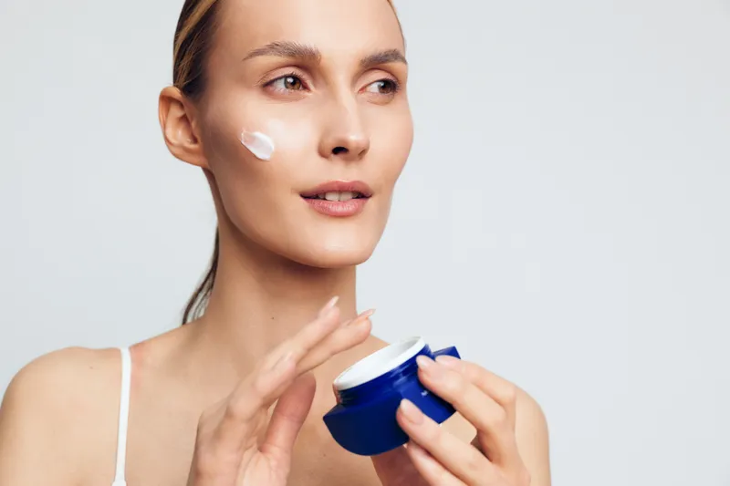 Young woman applying face cream on cheek, skincare routine, natural makeup, clear skin, soft lighting, white background, beauty concept, healthy lifestyle, closeup portrait