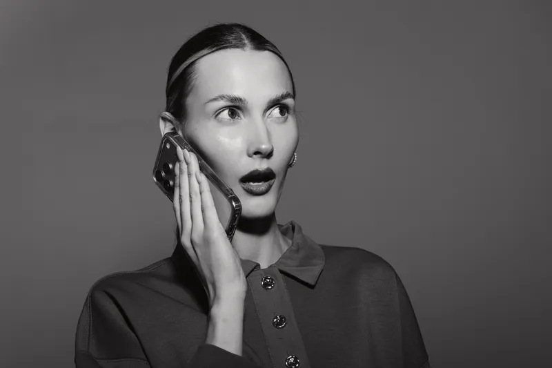 Young white woman with light skin talking on smartphone with surprised expression. Studio portrait in black and white. Concept of communication, emotions, and technology usage.