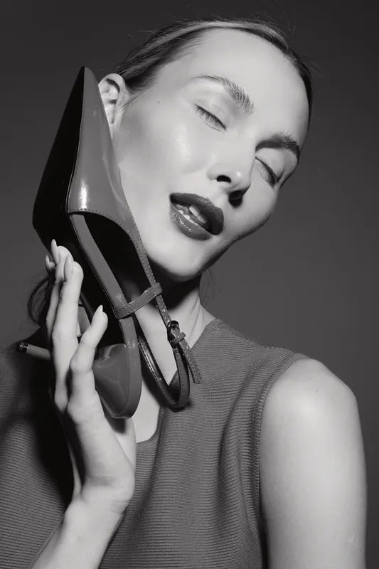 Young Caucasian woman with fair skin holds high heel shoe near face, eyes closed, wearing sleeveless top in black and white studio portrait.
