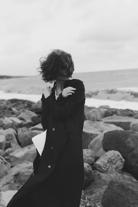 Woman in a long coat stands on a windy beach among rocks, facing the sea with a notebook tucked under her arm, as overcast light and shoreline haze create contemplation