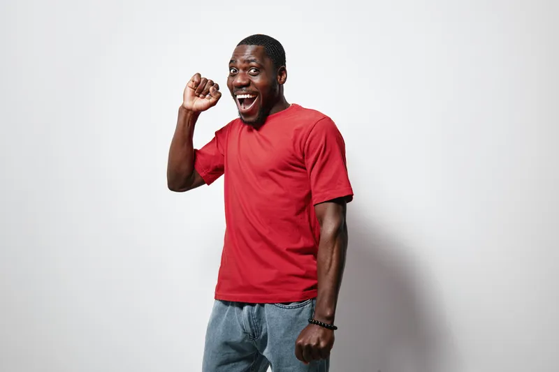 African American man with dark skin wearing red t shirt and jeans smiling and posing with raised fist on white background. Happy male model with casual style and joyful expression.