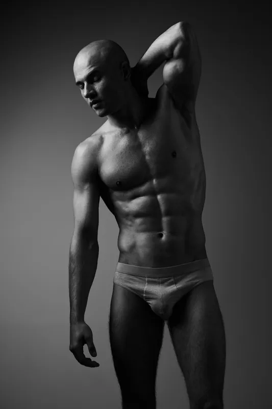 Black and white image of a muscular man posing confidently, showcasing strength and fitness, with dramatic lighting highlighting his physique.