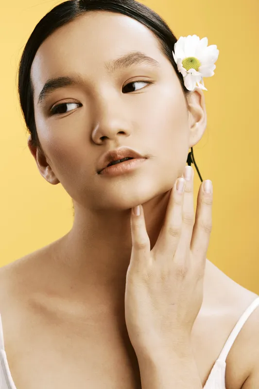 Asian woman with a white flower tucked behind her ear, poised against a warm yellow backdrop, radiates calm elegance and refined beauty in a serene studio portrait.