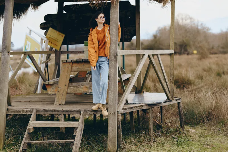 Woman lifestyle outdoors countryside field wooden platform rustic portrait daylight, standing full body at worksite, wearing brown jacket and jeans, calm confident fashion look in natural light