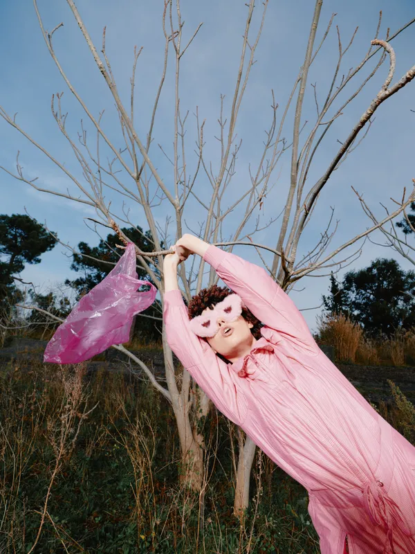 Woman in a pink outfit reaches for a bag while hanging from a bare tree in an outdoor landscape, expressing playful fashion and a carefree spring mood.