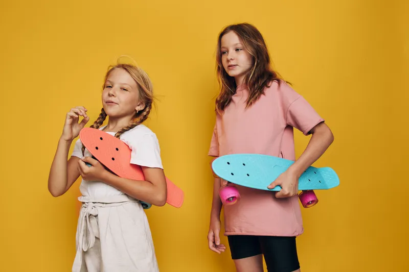 Two girls holding colorful skateboards pose on yellow background. One girl wears pink shirt, other girl wears white top and shorts. Studio portrait.