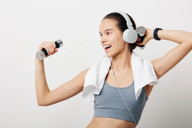 Woman fitness enthusiast in a workout scene with dumbbells, headphones and towel, smiling mid exercise, showcasing active gym energy and focus.