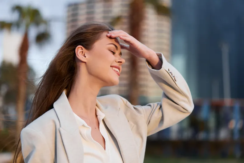Smiling woman in beige suit with long hair enjoying sunshine in urban outdoor setting, expressing happiness and confidence in modern business style