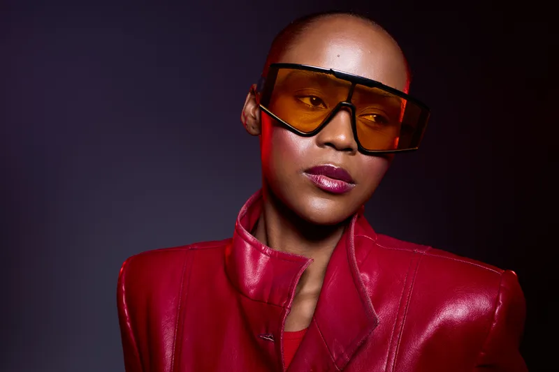 Stylish bald African woman with dark skin wearing oversized orange sunglasses and a red leather jacket poses against dark background. Concept of fashion, confidence, and modern style.