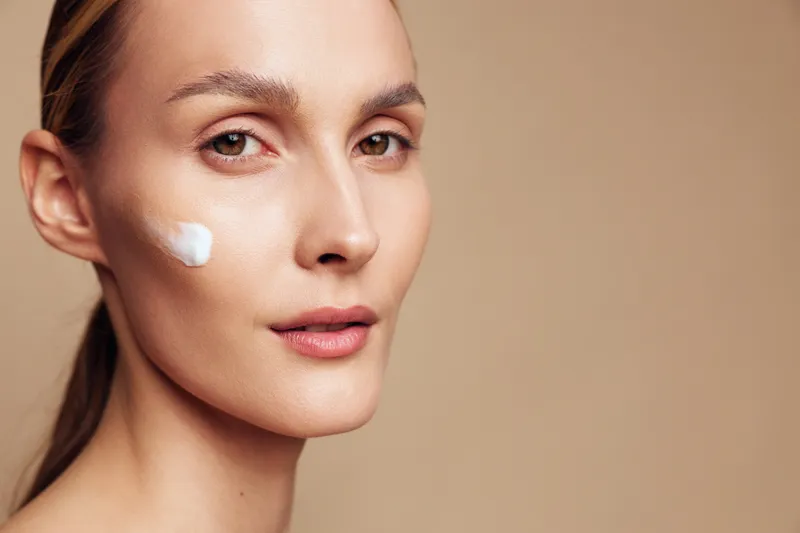 Young woman with natural skin applies moisturizing cream on cheek looks at camera with calm expression isolated on beige background skincare beauty concept.