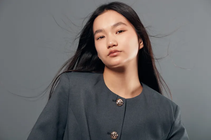 Young woman portrait with long black hair wearing buttoned gray jacket standing on gray background. Calm expression and natural makeup with elegant outfit and studio lighting.