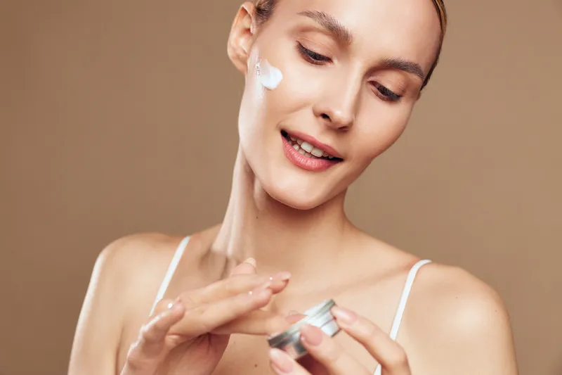 Young woman applying white face cream on cheek with fingers. Close-up portrait on brown background. Concept of skincare, beauty, wellness, and self-care routine with soft lighting.
