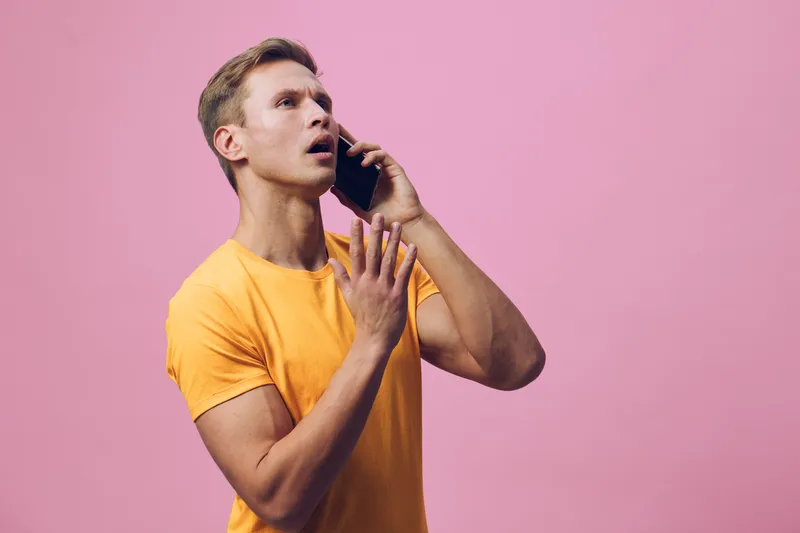 Confused young man with blond hair wearing orange T-shirt talking on smartphone isolated on pink background. Portrait of a surprised man reacting to unexpected news or situation. People lifestyle conc