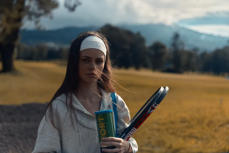 Young female athlete holding tennis rackets and a drink, showcasing sporty attire and determination in a picturesque outdoor setting