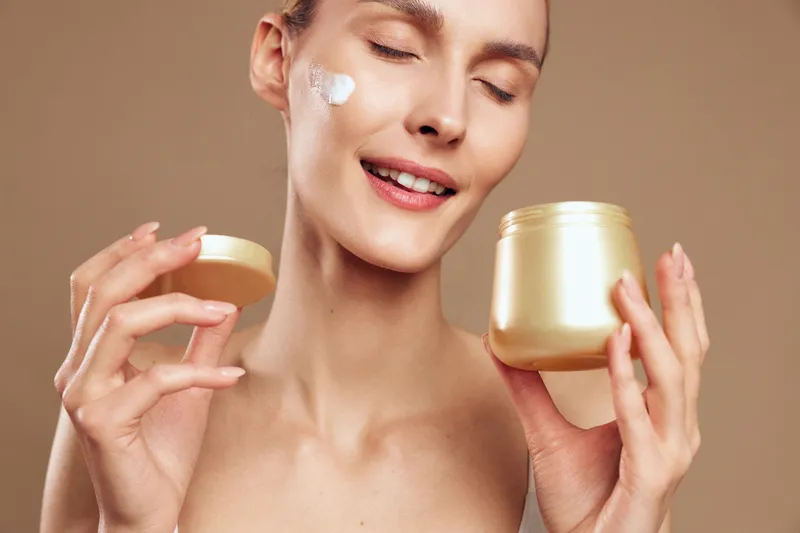 A young woman with clear skin applies moisturizing cream on her face while holding an open jar. She smiles with closed eyes against a plain beige background, skincare concept.