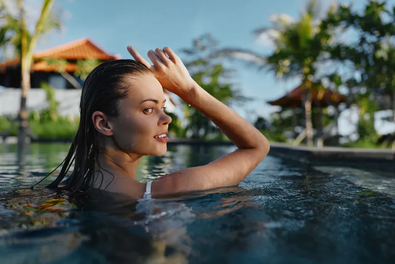 A woman with wet hair rises from a sunlit pool, smiling softly as she enjoys a peaceful, carefree moment in warm weather.