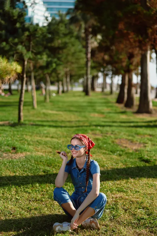 smiling girl sitting on grass wearing sunglasses and denim jumpsuit with red headband enjoying sunny day in park with trees and green lawn around