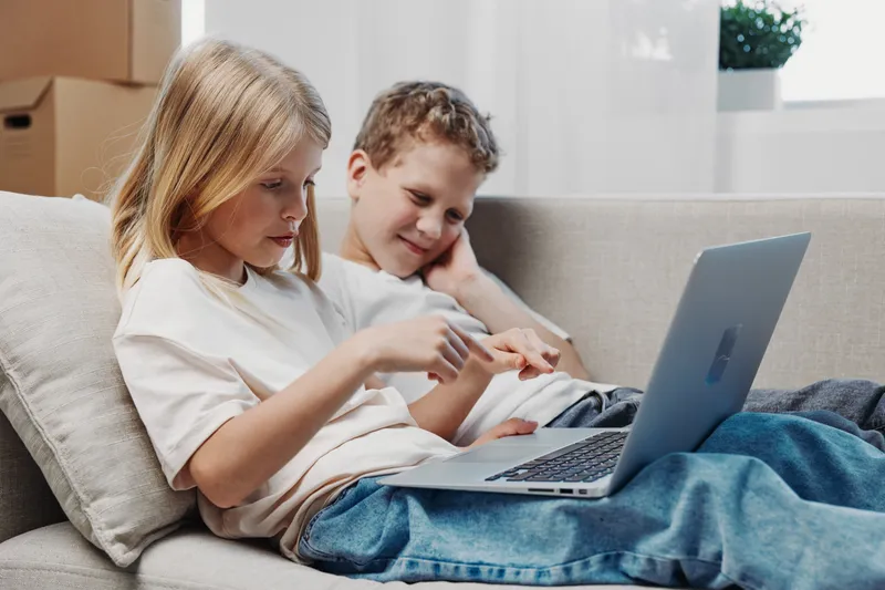 Two children sitting on a couch using a laptop, focused on the screen in a bright living room. Concept of technology, learning, and casual family time at home.