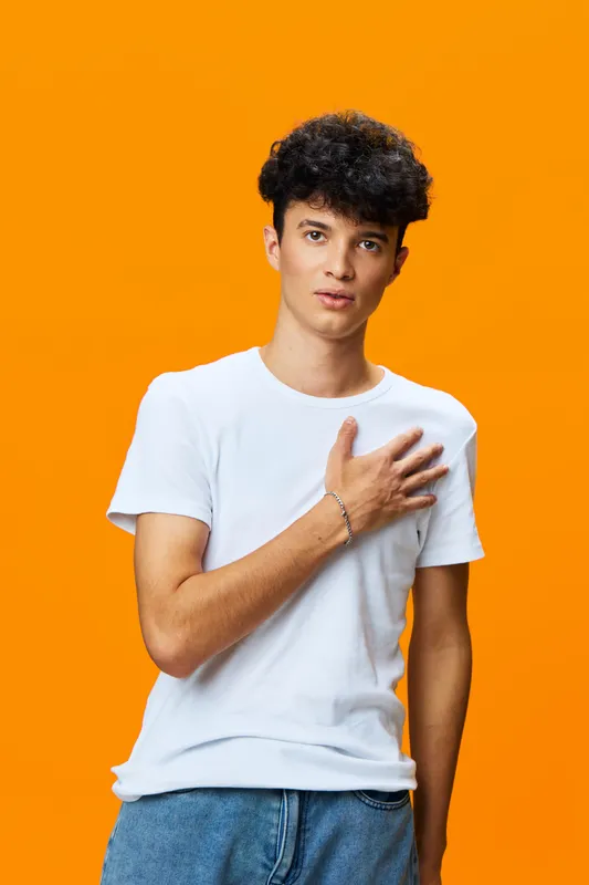 Young man with curly hair wearing white t-shirt and blue jeans standing against vibrant orange background with hand on chest, showing sincerity and calm expression.