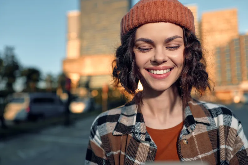 Woman smiling in urban portrait in a beanie and plaid jacket, sunlight and golden hour glow creating authentic candid lifestyle scene, emotional storytelling and mindful living.